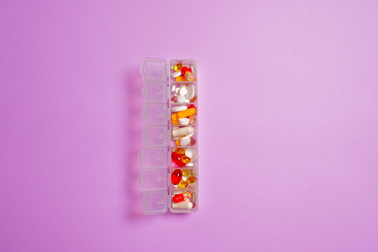 hero-img-02 Close-up of assorted pills in a weekly organizer against a violet background.